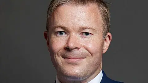 UK Parliament A posed shot in front of a grey background of a man smiling. The collar of a white shirt and navy jacket is just seen. He has brown short hair and brown eyes.