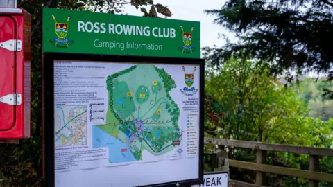 A large green information board for Ross Rowing Club stands beside a wooden footbridge, surrounded by trees and greenery.