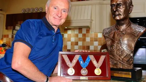 Brian Reid A man with white hair looks into the camera. He is wearing a royal blue top with short sleeves. He is holding a red box frame containing three medals. A bust of himself is also beside him.