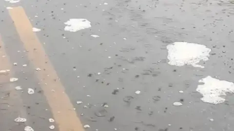 Puddle of water with soapy looking suds on road with two yellow lines.