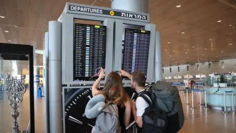 Travellers, carrying rucksacks, point at a flight information board in the departure lounge of an Israeli airport