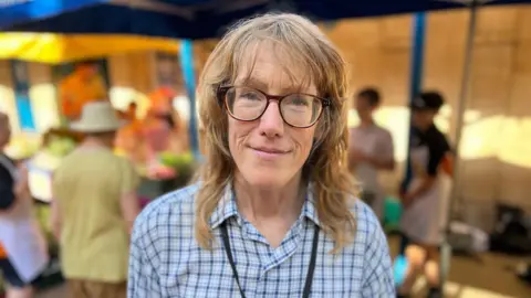 A person wearing glasses and a checked shirt, stands outdoors in front of market stalls. A lanyard hangs around their neck with canopies, tables and several people visible in the background. 