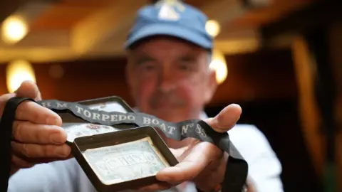 John Fairhall/BBC Alan Coday holds a Franc note and black torpedo boat ribbon aloft to the camera. He is out of focus in the background. He is standing in a dimly lit room below deck.
