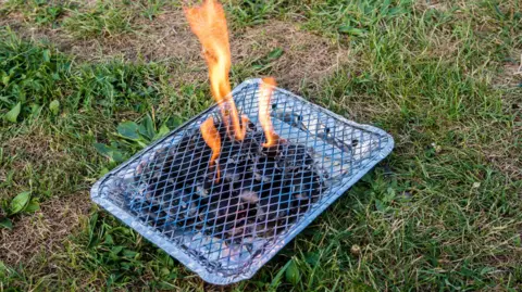 A stock image of a lit disposable barbecue, showing flames emerging through a grill on a foil case.