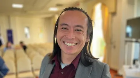 A man with shoulder length black hair held back by a headband, with a grey blazer and a maroon shirt, smiles at the camera. Behind him there is a function room with chairs lined up. 