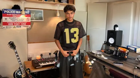 BBC Kamal stands in a studio surrounded by guitars, a laptop, keyboard and speakers. He is holding on to the belt of his black trousers and wears a black top with the number 13 embossed on it in yellow. 
