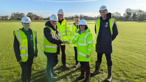 Six people stand in a green field, wearing white hard hats and smile at the camera 