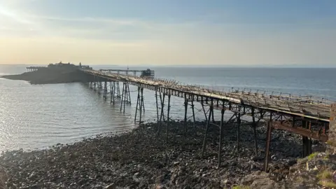 A view of Birnbeck Pier in Weston-super-Mare taken from the shore with the rocky and muddy beach in the foreground and the crumbling, rusty pier going out to see with the ruins on Birnbeck Pier visible in the distance. It is a sunny day and appears to be early in the morning