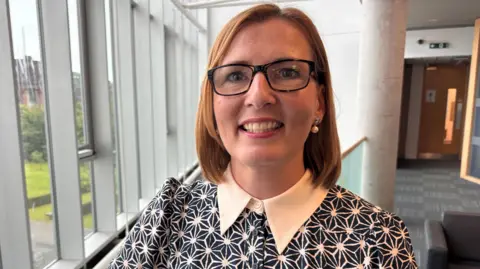 Donna Stewart has shoulder-length red hair and black glasses. She is in a corridor and is smiling. She is wearing a black and white patterned top with a pointy white collar.