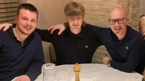 Harry, Freddie and Alain sat with their arms round each other at a table in a restaurant, with glasses on the table and a salt or pepper shaker.