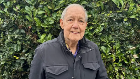 Paul is standing in front of a green bush. He is wearing a charcoal coloured jacket. He is smiling slightly.