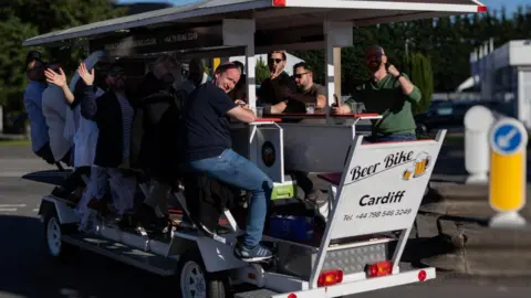 A group of men are smiling while sitting on a beer bike which says 'Beer Bike Cardiff'. They each have a plastic cup on a table in front of them and a set of pedals and there is a white canopy over their heads.