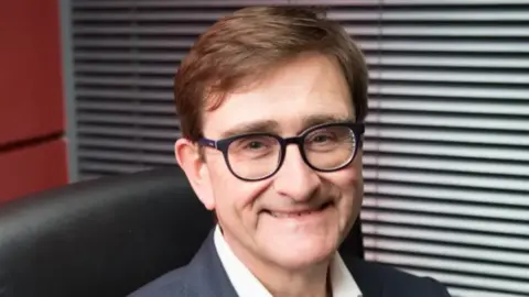 A man with glasses and brown hair sat in an office chair smiling into the camera.