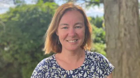 Lewis Adams/BBC Marie Goldman is standing outside in front of a tree. She is looking directly at the camera and smiling and is wearing a navy patterned short sleeved T-shirt.