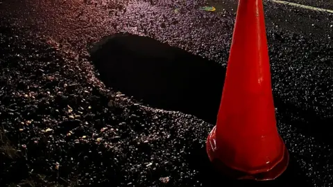 Becky Broadley A large pothole on a road, in the dark, with a red cone by it. 
