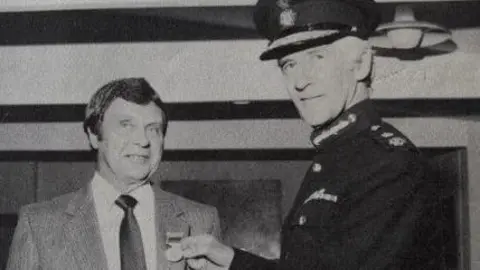 Harwell Campus A black and white photo of a man in a suit being presented with a medal by a taller, older man wearing offical Lord Lieutentant military style dress. Both are smiling and the Lord Lieutenant holds a British Empire Medal onto Griff Bowen's chest.