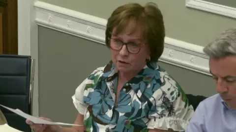 Diane Dodds has short reddish-brown hair and glasses and is wearing a floral blouse. She is holding a letter in her hand