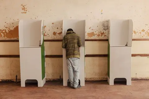 AFP via Getty Images A voter at a cardboard ballot booth has their back to the camera. The voter is in the middle of three ballot booths set against a scuffed wall. 