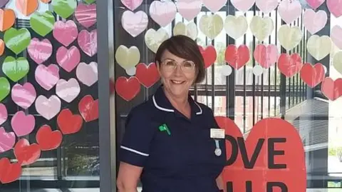 Carol Wilkins Carol is wearing a dark blue uniform with white piping and a name badge. She stands in front of a glass wall decorated with colourful paper hearts. The hearts are arranged in rows and include shades of red, pink, green, and gold.