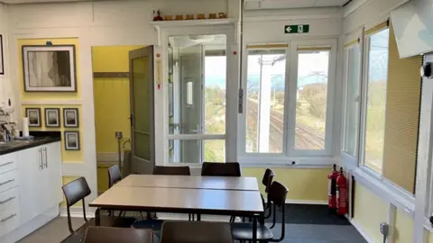Network Rail The inside of the signal box which is set up like a classroom with tables and chairs in the middle, white kitchen units to the left by a door to a cloakroom with yellow walls. There are large white framed windows with a view of the railway track