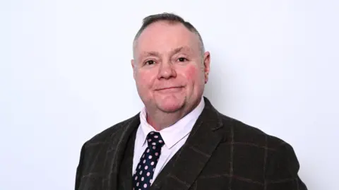 Labour Party A man with short dark hair in a dark suit with a dark blue patterned tie in front of a white background