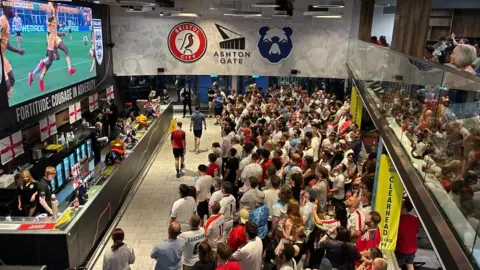 Isobel Gauld A crowd of fans watch the England match on a big screen at Ashton Gate next to a bar and wall with the Bristol City FC and Bristol Bears logos.