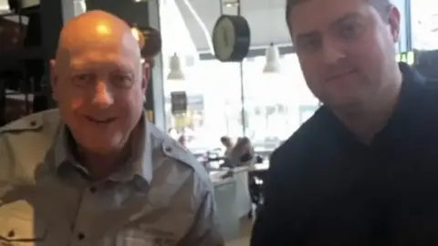 A photo of two men - one wearing a grey shirt and one wearing a dark shirt. They are sitting in a restaurant and looking at the camera. 