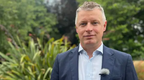 Luke Pollard with short grey hair, wearing a blue jacket and shirt standing in a garden