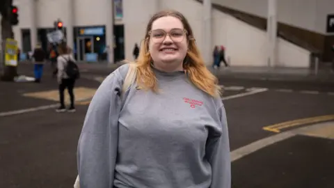 A smiling girl in a grey jumper stood with her bag on her shoulder. She wears transparent glasses. In the background is a Greggs and several pedestrians.