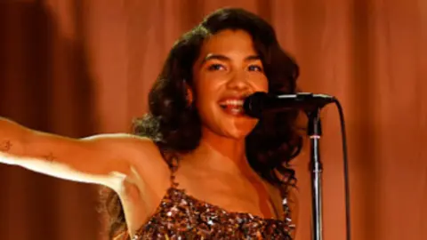 Dean with long, wavy dark hair wearing a sparkly flapper dress, singing into a microphone with one arm stretched out to the side, pointing. Behind her is a gold curtain.