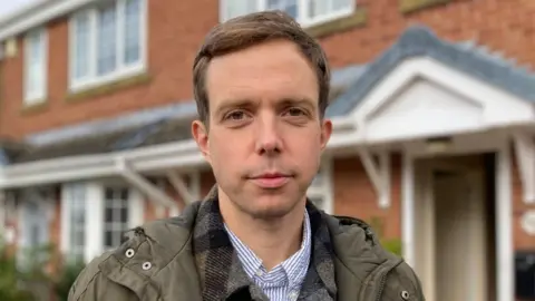 A man with short brown hair looks into the camera. He is wearing a striped blue shirt, a grey plaid shirt and a green coat. He is standing outside a house with red bricks and white windows and doors.