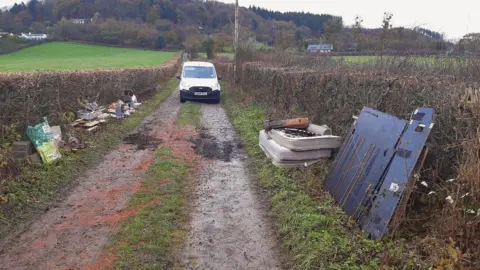A country lane is pictured with mattresses, building waste and metal sheets piled on the grass verges on either side. A white van is parked on the road in shot.