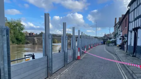 Environment Agency A road with houses running down one side and metal posts on the other, with slats running between them to form a wall, with the river on the other side and a bridge in the distance