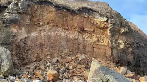 BBC / Glenn Pringle An exposed cliff face with rocks on the ground.