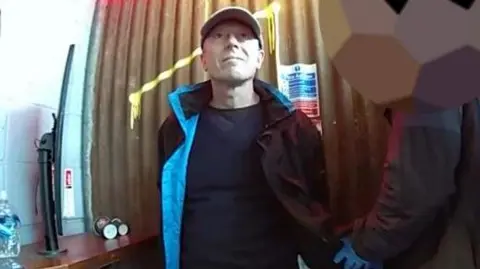 Met Police Howard Phillips wearing a jacket and cap while being arrested by a police officer, who is blurred.
