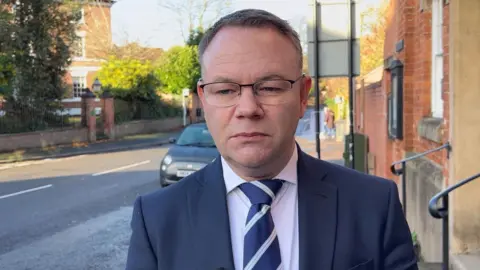 Philip White wears glasses, a suit and tie, is standing on a street in Tutbury. He has a serious expression. 