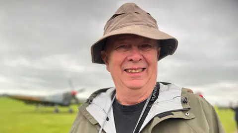 Emma Baugh/BBC Donald Johnson wearing a beige hat and coat with the airfield and an aircraft behind him, Duxford