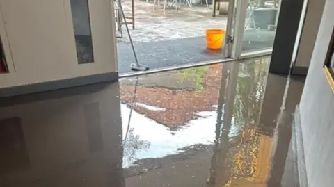 A flooded entrance space to an art gallery. There is water all over the floor and, in the doorway to outside the property, there is a broom and an orange bucket. There is an outdoor seating area.