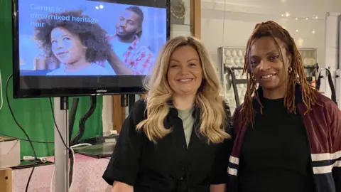 A woman with long blonde hair and a woman with braided brown afro hair are smiling as they stand next to each other. An image on a screen behind them says 'caring for afro/mixed heritage hair' with an image of a man and a young girl, both with afro hair.