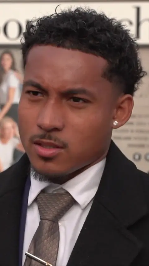A man with short curly hair looks off camera with a furrowed brow. He has a moustache, some facial hair on his chin, dark curly hair on his head and an ear stud. He is wearing a white shirt buttoned up with a tie and a black jacket.