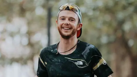 David Altabev Will Robinson wearing a black running t-shirt and a white cap backwards. He has short brown hair and a ginger beard. He is wearing reflective sunglasses on his head and smiling at the camera as he runs through a park. In the background there are trees which are out of focus.