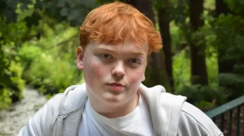 Mr Horne is wearing a white top and light jackets while standing in font of trees with lots of green leaves. He is looking directly at the camera and has ginger short hair.