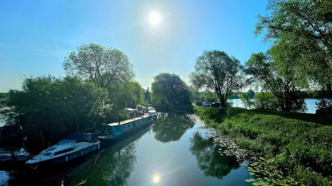 Weather Watchers/Alfie Cole The sun is reflected as a perfect yellow circle in the middle of a canal, with small boats and narrow boats parked on one side. 