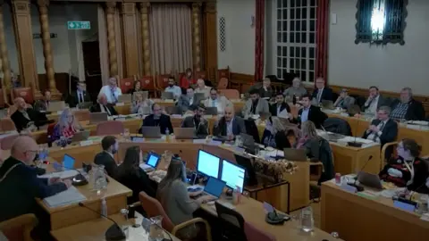 Peterborough City Council Council meeting in progress- with councillors sitting on wooden tables in a semi-circle with the mayor sitting in front alongside council officers.