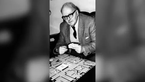 A black and white photo of an old man wearing glasses and a suit, playing Cluedo