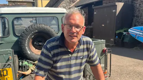 Simon is wearing a blue and green striped polo shirt and glasses. He is sat on the back of a truck in a farm yard.