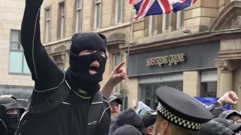 A masked individual in dark clothing raises a clenched fist while standing close to a police officer in a high-visibility jacket and checkered hat, with a crowd gathered behind and a Union Jack flag visible above on a city street lined with stone buildings and shopfront signage reading “Sostrene Grene.”