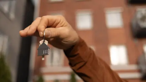 Getty Images A hand holds up a key and a keyring in the shape of a house. A block of flats is in the background.