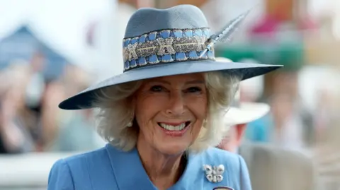 PA Close-up portrait of a person wearing a light blue jacket and a wide-brimmed blue hat decorated with patterned ribbon and a feather. A butterfly-shaped brooch is pinned to the lapel of the jacket. The background is softly blurred, showing an outdoor event setting with indistinct figures, tents, and seating. Bright natural light illuminates the scene, highlighting the textures of the hat and clothing. No clearly readable text is visible in the image.