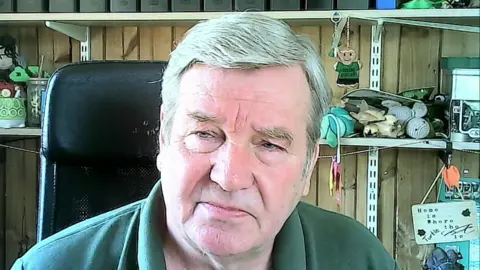 A man sits in a desk chair within an office. He has grey hair and is wearing a green polo shirt. There are shelves behind him that have various items sitting on them.